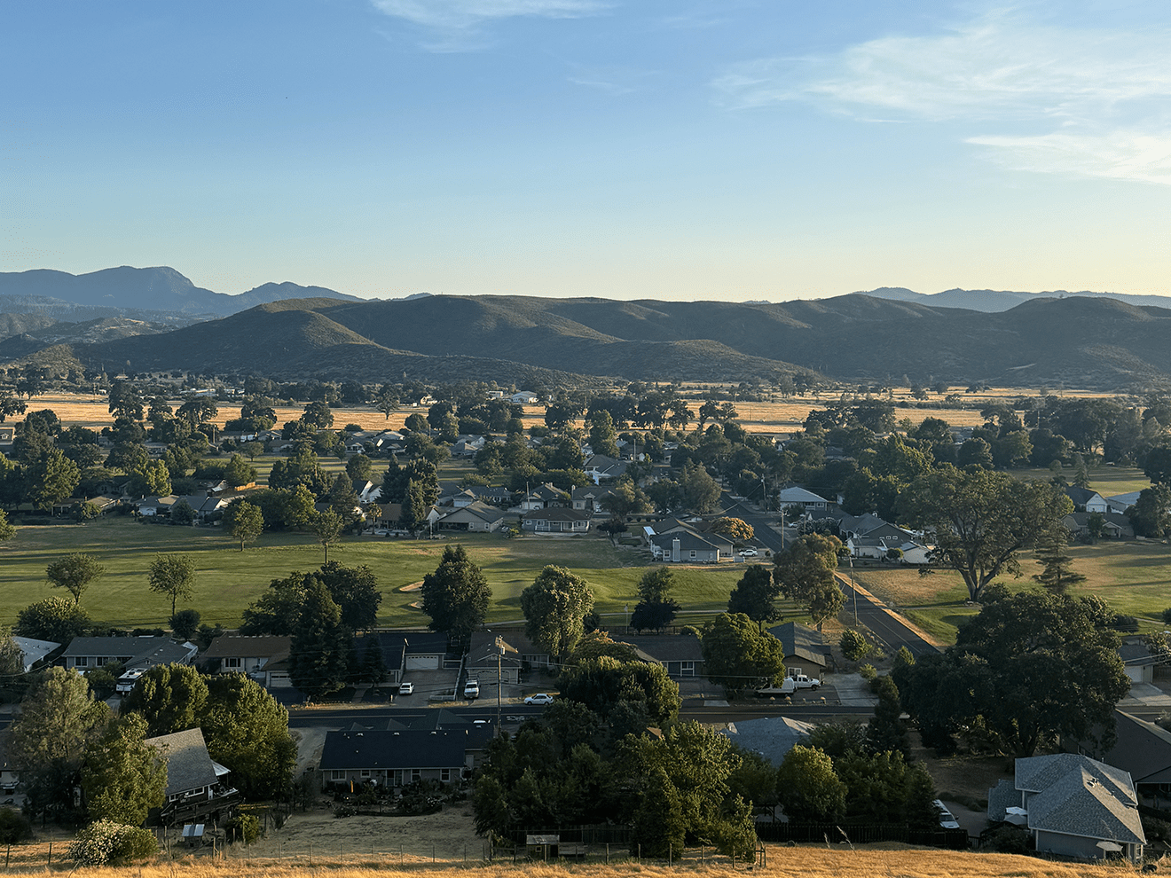 A scenic view of Hidden Valley Lake, CA, showcasing a peaceful residential neighborhood surrounded by rolling hills, green golf course fairways, and golden summer grasslands under a clear blue sky.